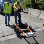 Two workers use a portable ground penetrating radar system on a roadway.