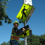 Worker installing a flashing beacon on a pedestrian crossing sign.