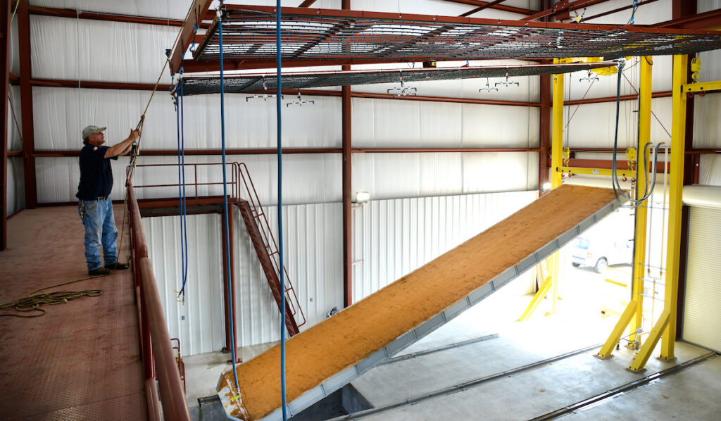 A man stands on a metal platform inside an industrial building next to a sloped conveyor or ramp leading to yellow support beams.
