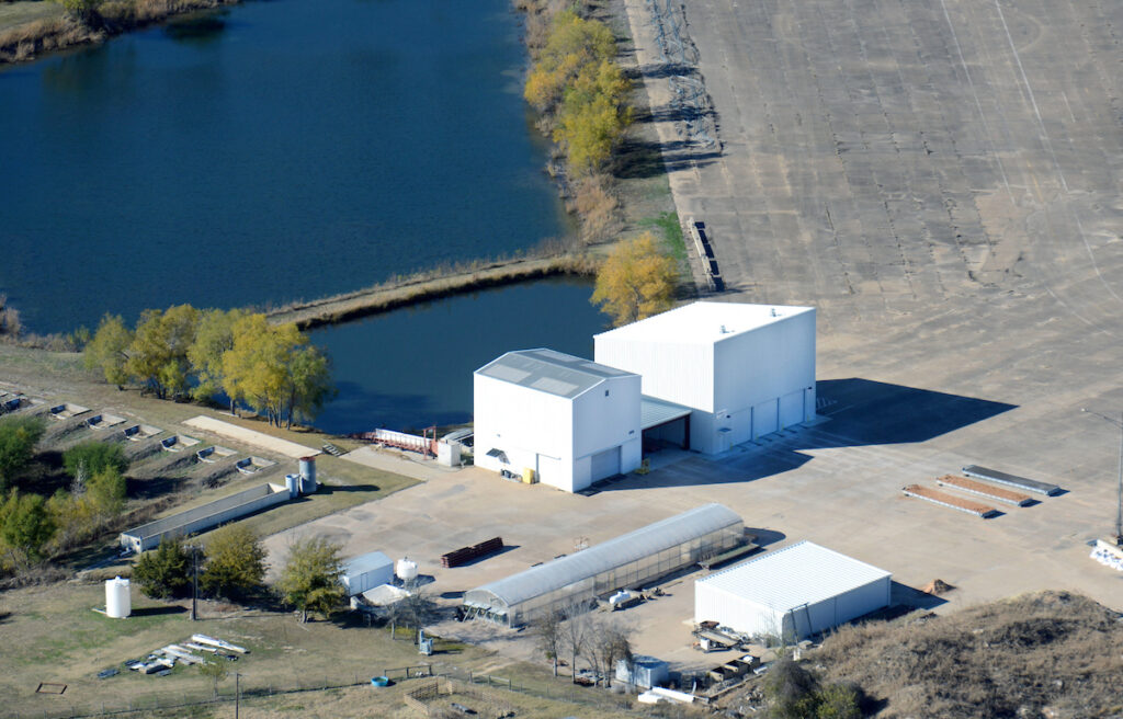 Aerial view of an industrial facility with several white buildings, paved areas, and adjacent water features surrounded by trees.