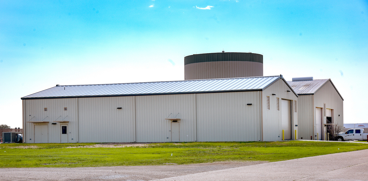 A large, beige industrial building with a metal roof sits on a grass lot under a clear sky; a tall, cylindrical structure is behind it, and a white truck is parked nearby.