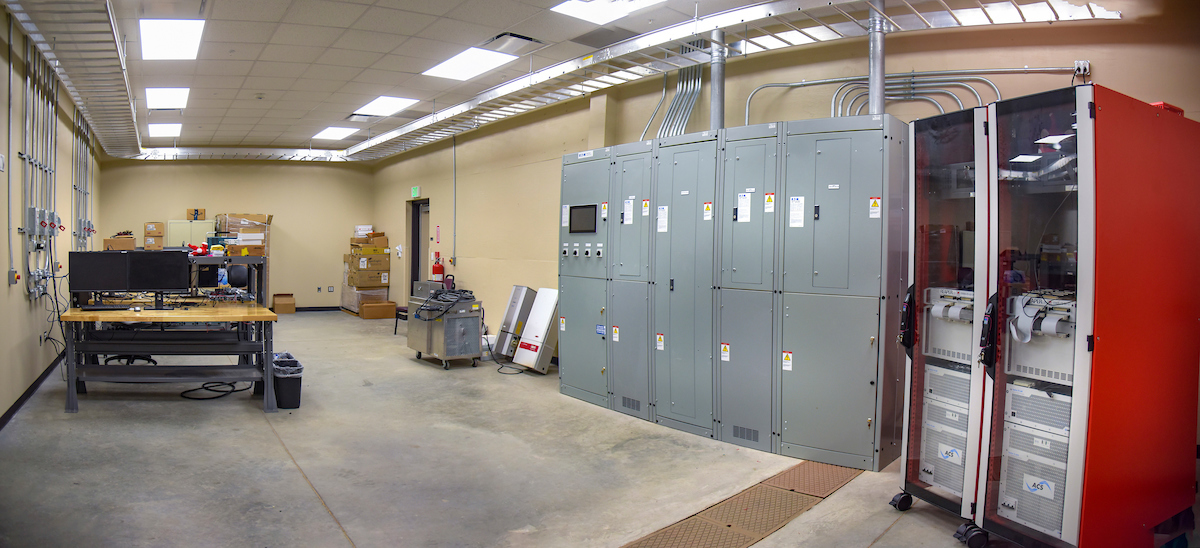 Room with electrical control panels, server cabinets, and workstations; boxes and equipment are arranged along the walls under fluorescent lighting.