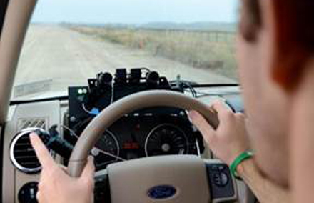 Person driving a Ford vehicle on a dirt road, with hands on the steering wheel and electronic equipment mounted on the dashboard.