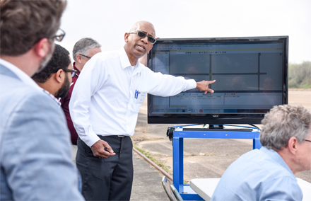 A man in a white shirt points at a diagram on a large monitor while others stand around him outdoors.