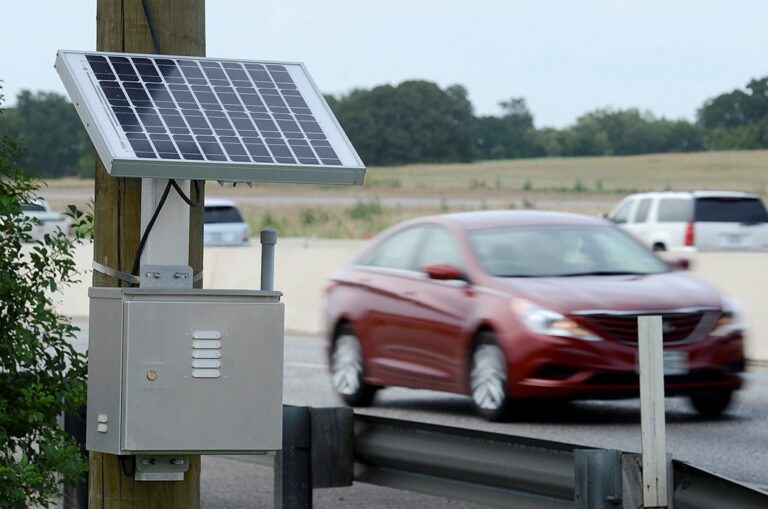 A roadside solar panel installation is mounted on a pole next to a highway, with cars driving by in the background.