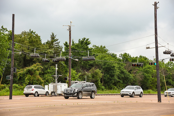 Several cars wait at a large, multi-lane intersection with overhead traffic lights and wires on a cloudy day, surrounded by trees.