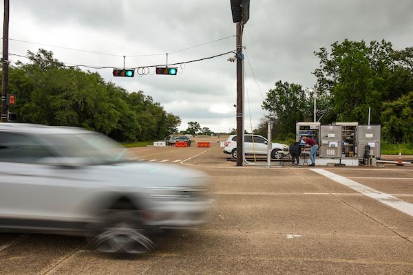 A blurred car drives through a green light at an intersection, while two people use gas pumps at a nearby station under a cloudy sky.