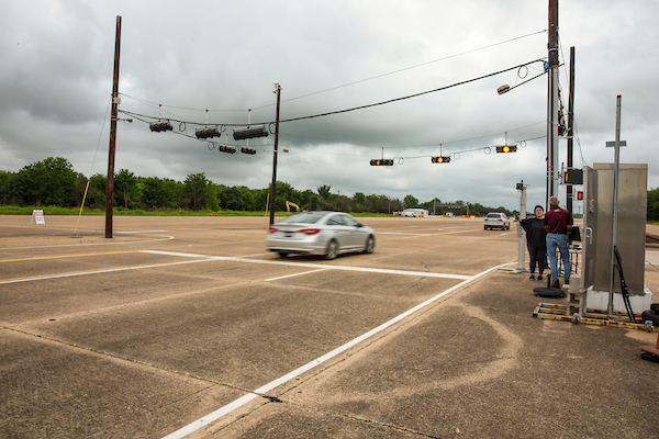 A silver car speeds through an empty intersection with traffic signals, while a small group of people stand near equipment on the roadside under cloudy skies.