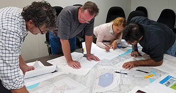 a group of people going over various kinds of roadway maps covering a large table
