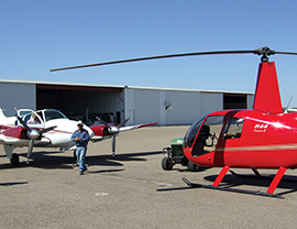 Small aircraft and helicopter parked outside of a hangar.