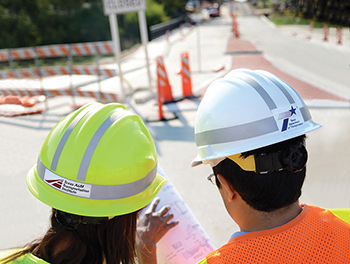 A person with a TTI hard hat and a person with a TxDOT hard hat looking at design plans at a road construction site.