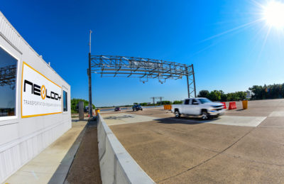 Exterior view of the Neology Transportation Research Center (TRC) facility next to the test track with vehicles approaching and passing under a test gantry.