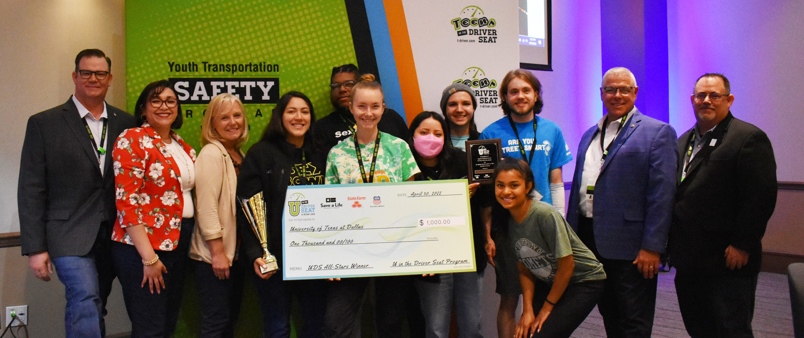 Group from The University of Texas at Dallas holding an All-Star winner check in front of a Youth Transportation Safety Program sign.