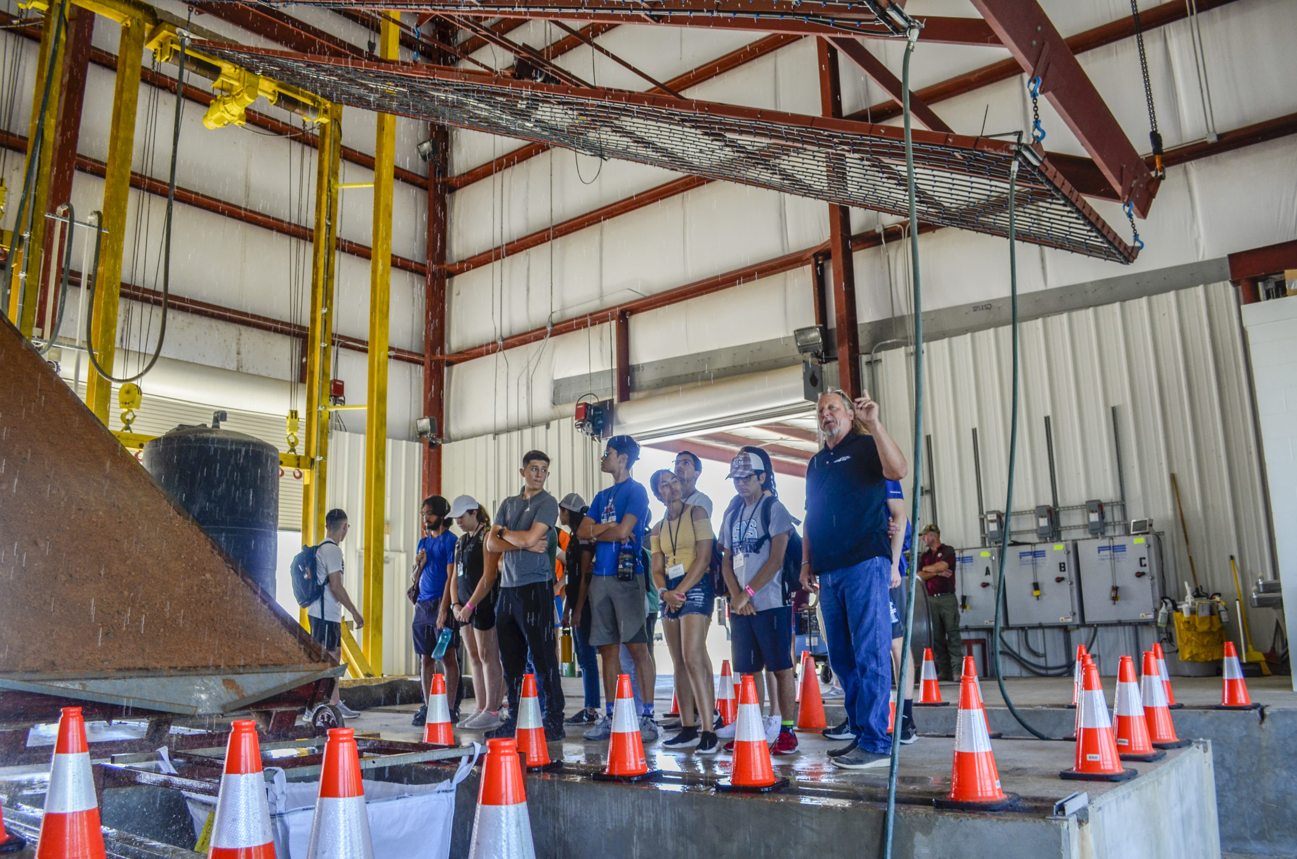 Photo of students and Jett McFalls inside a large building with a rainfall simulator above and a soil test bed in front of them.