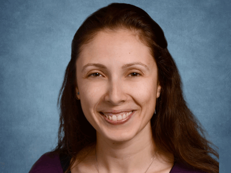 A woman with long brown hair and a purple top smiles at the camera against a blue textured background.