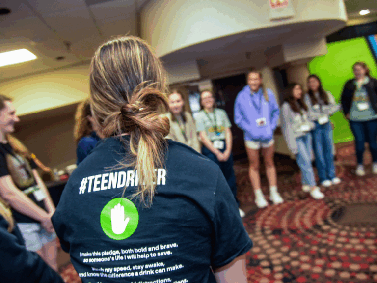A group of teenagers stands in a circle indoors; one person in the foreground wears a #TEENDRIVER shirt with a pledge written on the back.