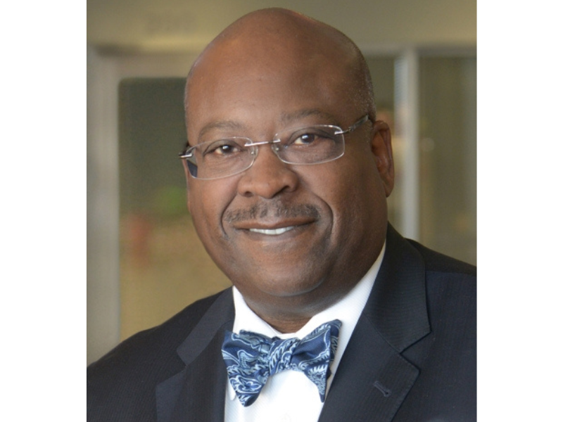 A man wearing glasses, a dark suit, a patterned blue bow tie, and a white shirt, smiling at the camera in an indoor setting.