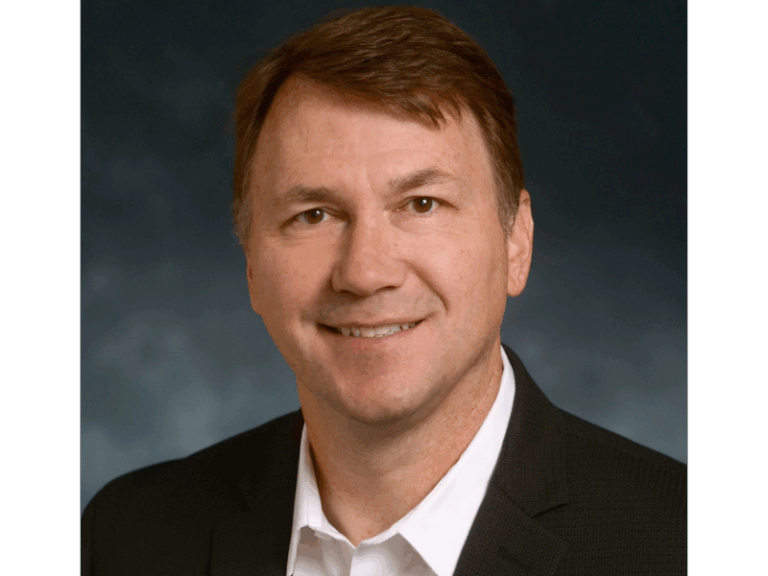A man with short brown hair wearing a black suit jacket and white collared shirt smiles at the camera against a dark, blurred background.