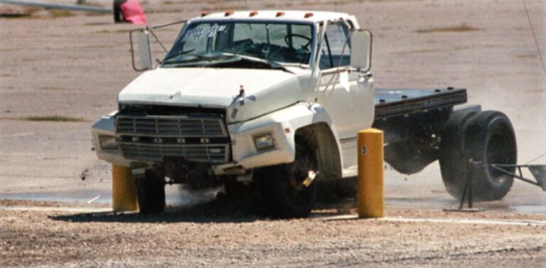 A white Ford truck collides with yellow bollards in a parking lot, causing visible damage to the front and raising dust.