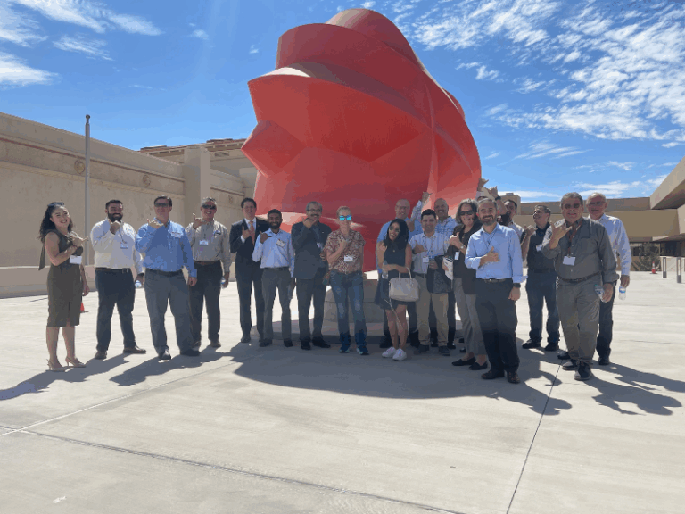 A group of people poses outdoors in front of a large red abstract sculpture on a sunny day.