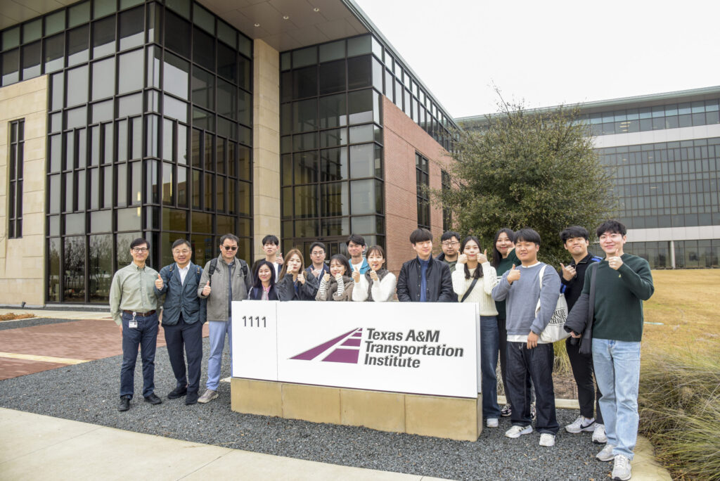 A group of people stands outside the Texas A&M Transportation Institute building, posing around a sign with the institute’s name and logo.