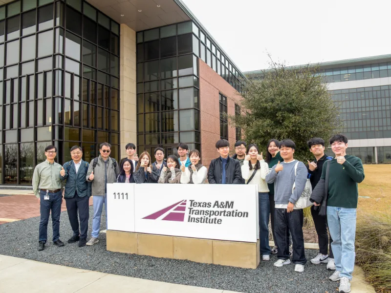 A group of people poses outside the Texas A&M Transportation Institute building, standing behind a white sign with the institutes logo.