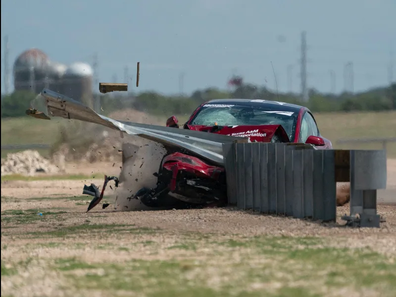 A red car crashes head-on into a metal guardrail, causing debris and damage to the front of the vehicle on a gravel area.