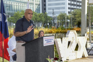 A man speaks at a podium labeled Investing in America outdoors, with V2X letters and city buildings in the background.
