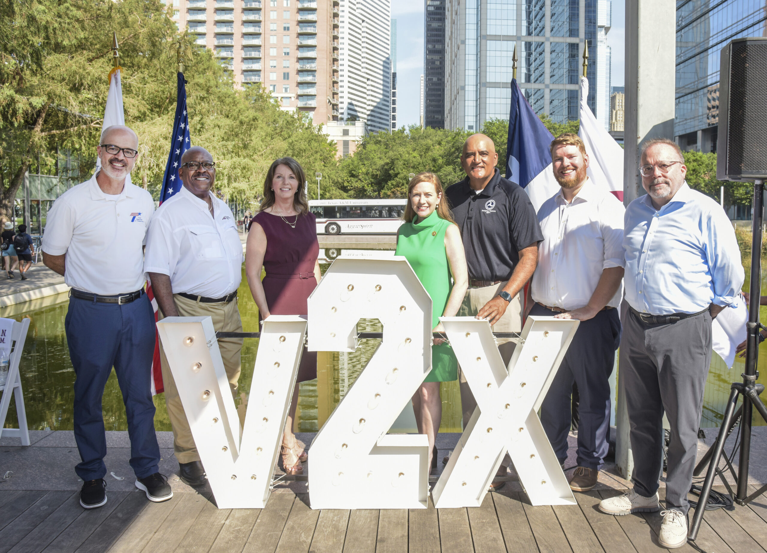 Seven people stand outdoors in front of large illuminated V2X letters, with city buildings, flags, and water in the background.