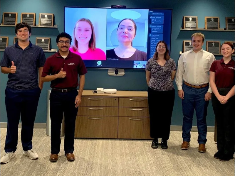 Six people, four standing in an office and two appearing on a large video screen, pose for a group photo in front of a wall with plaques.