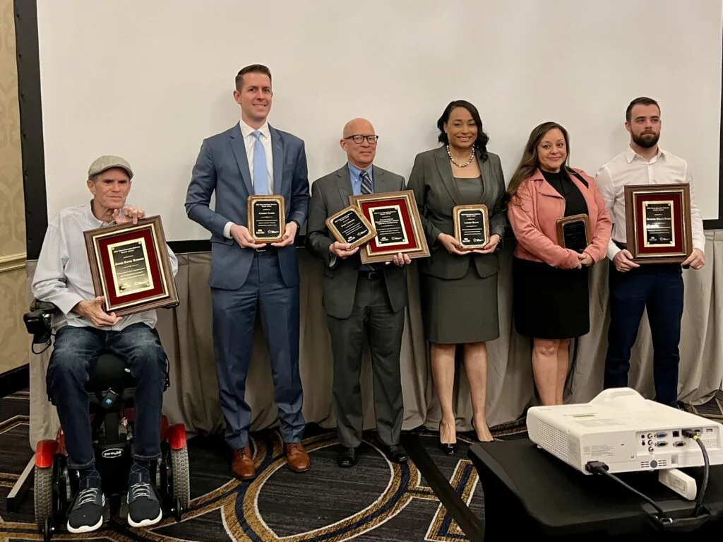 Six people stand in a row holding award plaques