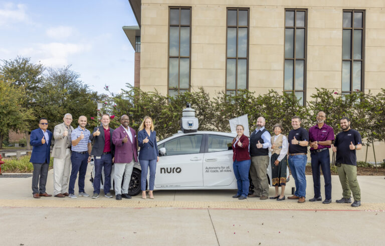 A group of people stand in front of a Nuro self-driving car, all giving thumbs up, with a building and trees in the background.