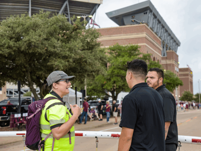 A woman in a yellow vest and gray cap talks to two men in black shirts outside a stadium, with trees and people in the background.