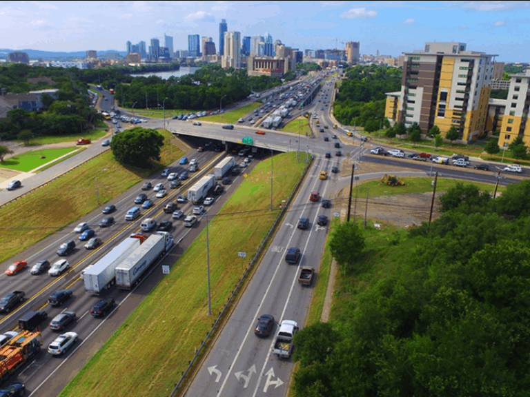 Aerial view of a busy highway interchange with heavy traffic, nearby buildings, and a city skyline in the background under a partly cloudy sky.