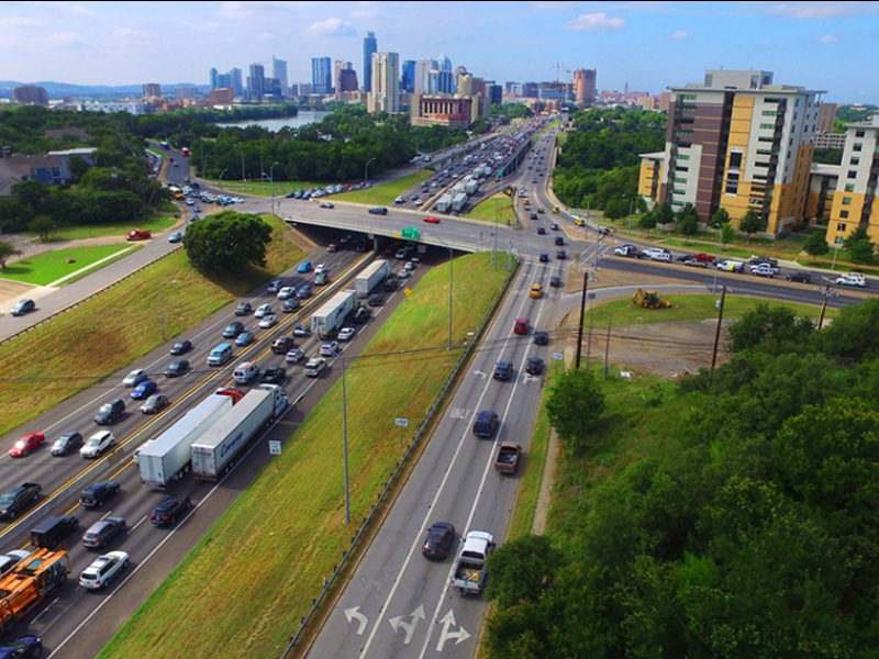 Aerial view of a busy highway interchange with heavy traffic, nearby buildings, and a city skyline in the background under a partly cloudy sky.