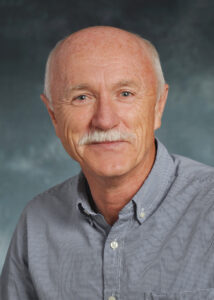 Professional headshot of Tom Scullion, wearing a checkered button-up shirt. He is smiling slightly and looking directly at the camera, with a neutral blue-gray gradient background.