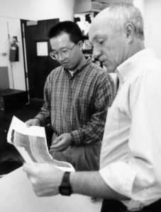 Black and white photograph of Yiqing Chen (left) and Tom Scullion (right) in the GPR Lab in the 1990s. Tom Scullion, wearing a striped button-up shirt, is holding a document while discussing its contents with Yiqing Chen, who is wearing glasses and a plaid shirt. They are standing in a laboratory setting with equipment and shelves in the background.