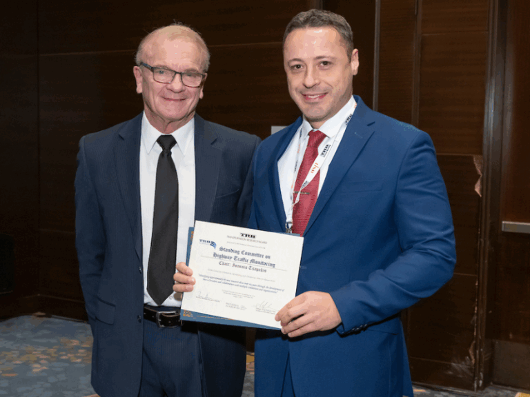 Yianni Tsapakis, wearing a blue suit and red tie, accepts the TRB Blue Ribbon Award for the Highway Traffic Monitoring Committee from another man in a dark suit and glasses. They are smiling and standing indoors at a formal event.