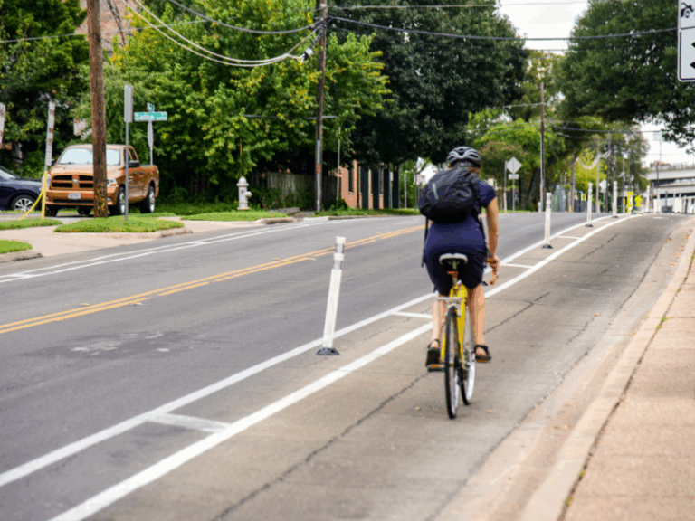 A person wearing a helmet rides a bicycle in a designated bike lane on a city street with trees and parked vehicles nearby.