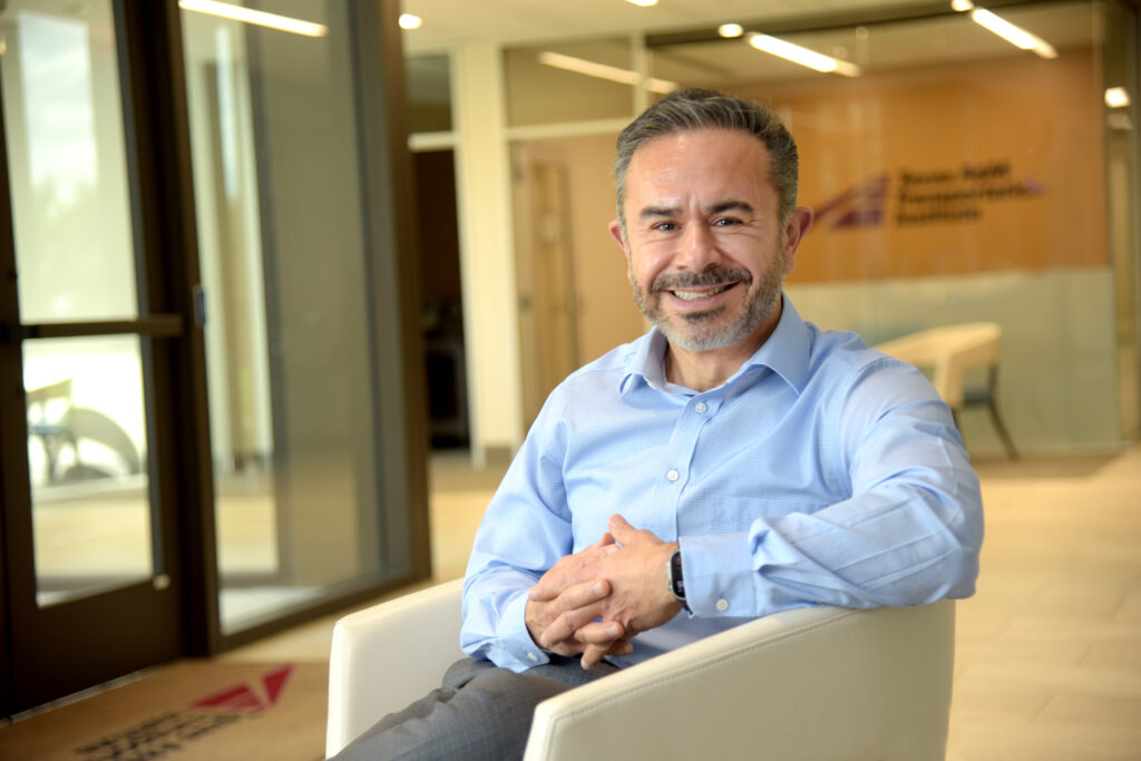 A man with short gray hair and a trimmed beard smiles while sitting in a white chair in a modern office lobby, wearing a light blue shirt. A glass door and a sign on the wall are visible in the background.