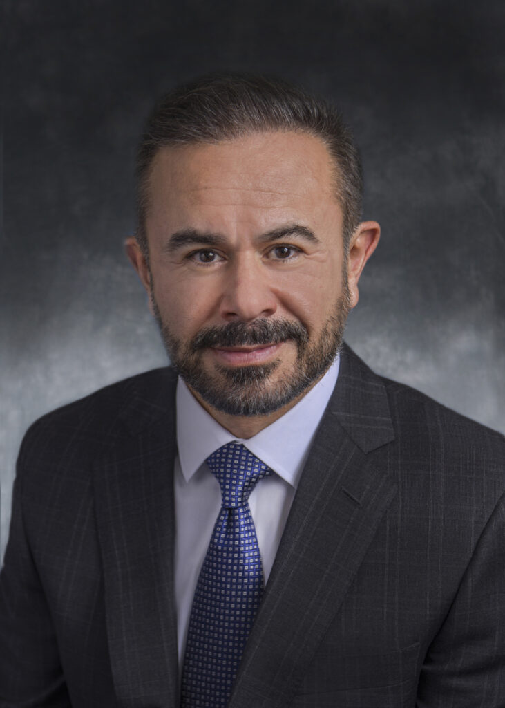 A man with short dark hair and a beard wearing a dark suit, white shirt, and blue patterned tie poses against a dark, mottled background.