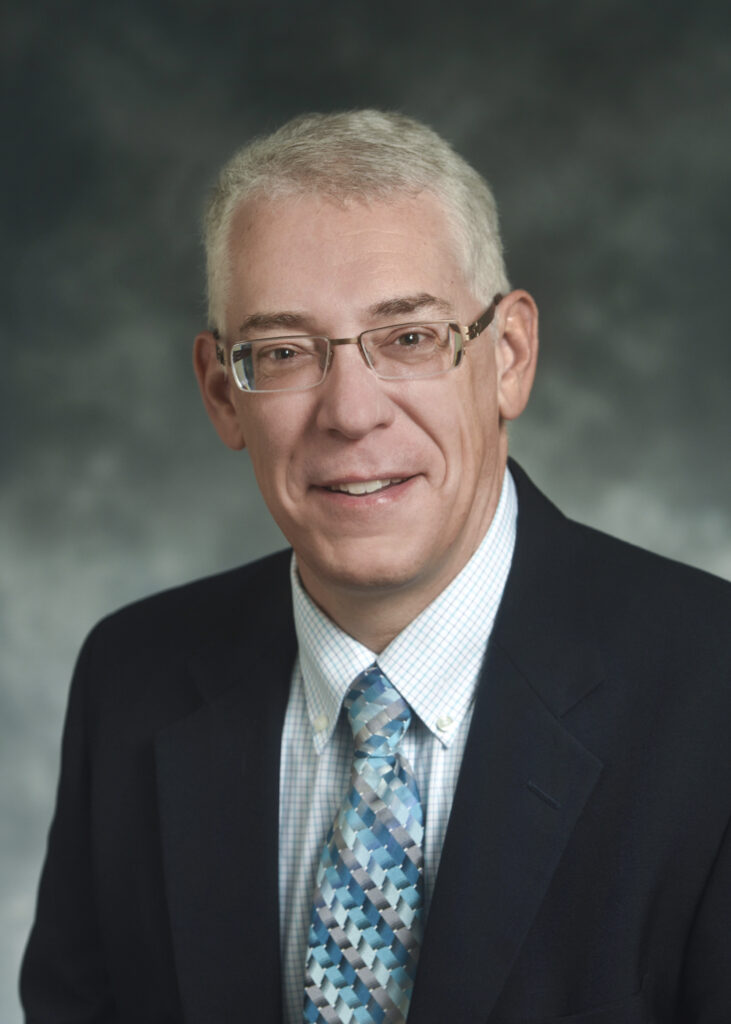 A middle-aged man with short gray hair, glasses, and a suit jacket poses for a professional headshot against a gray background.