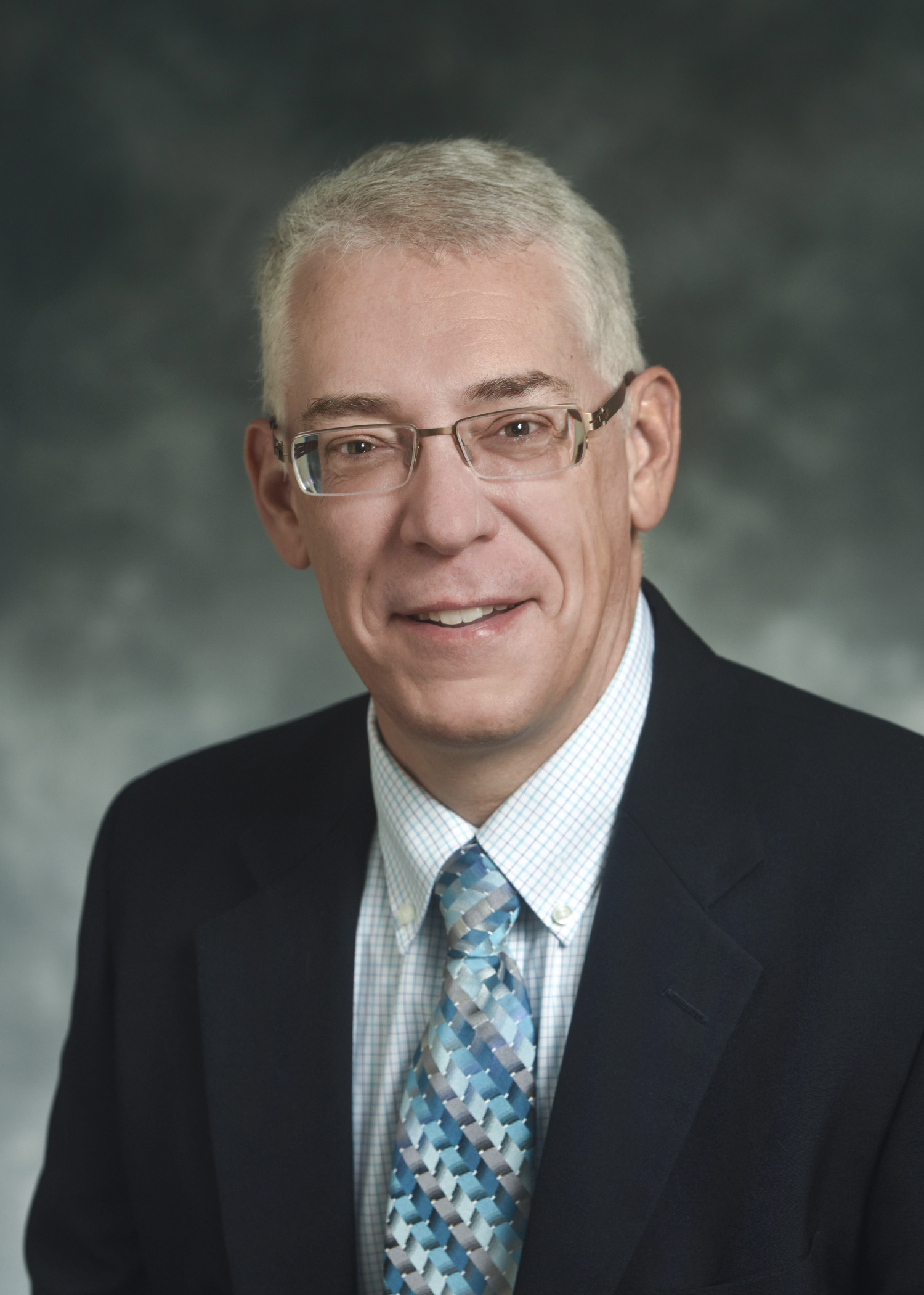 A middle-aged man with short gray hair, glasses, and a suit jacket poses for a professional headshot against a gray background.