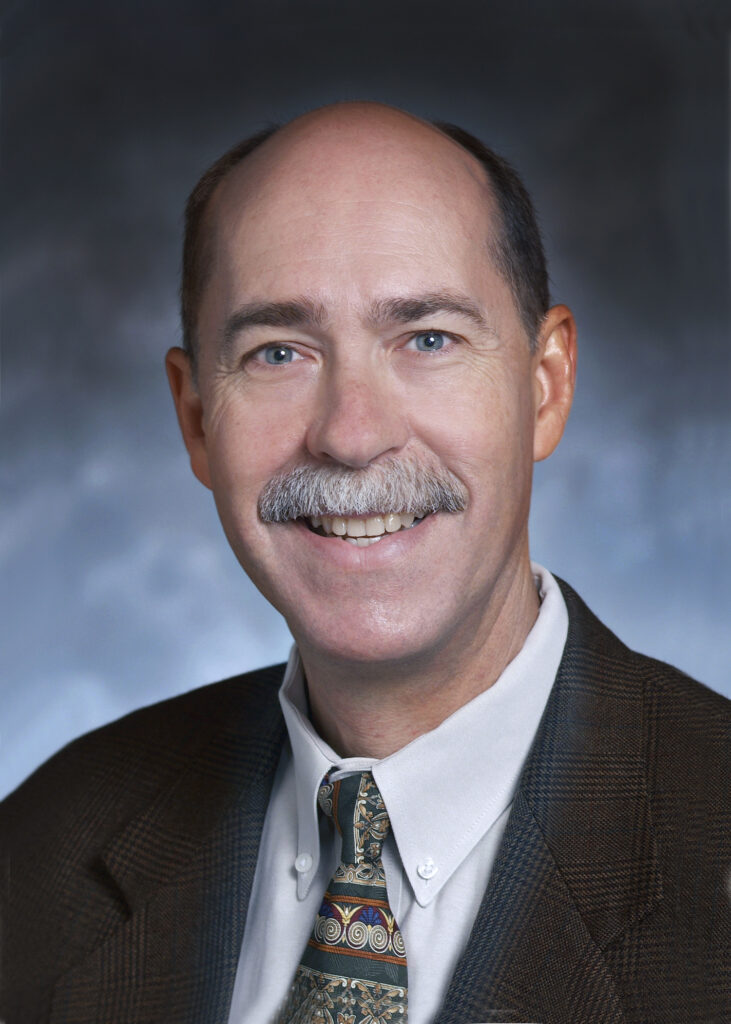 A middle-aged man with a mustache, wearing a brown suit jacket, patterned tie, and white shirt, smiling at the camera against a neutral background.