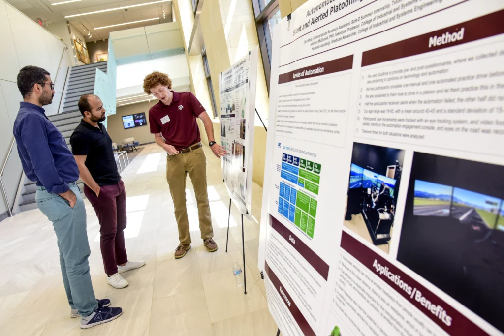 Three men stand in a modern building, with one presenting information displayed on large research posters to the other two.
