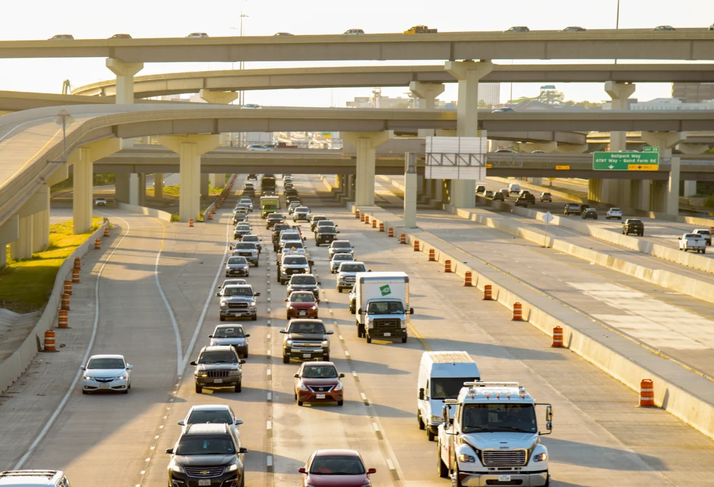 Multiple lanes of highway traffic move beneath elevated overpasses on a sunny day, with orange construction barrels lining the sides of the road.