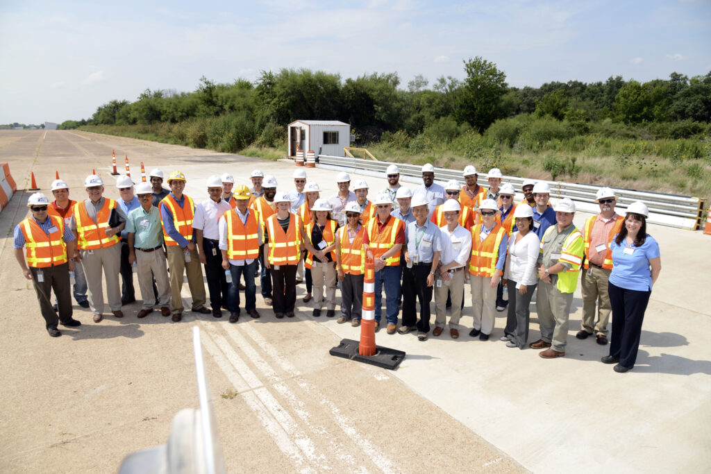 A group of people wearing safety vests and hard hats stand on a construction site outdoors, posing for a photo on a sunny day.