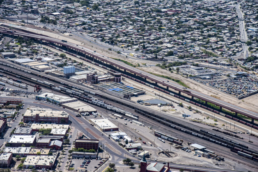 Aerial view of a rail yard with multiple train tracks, freight trains, industrial buildings, and a surrounding urban residential area.