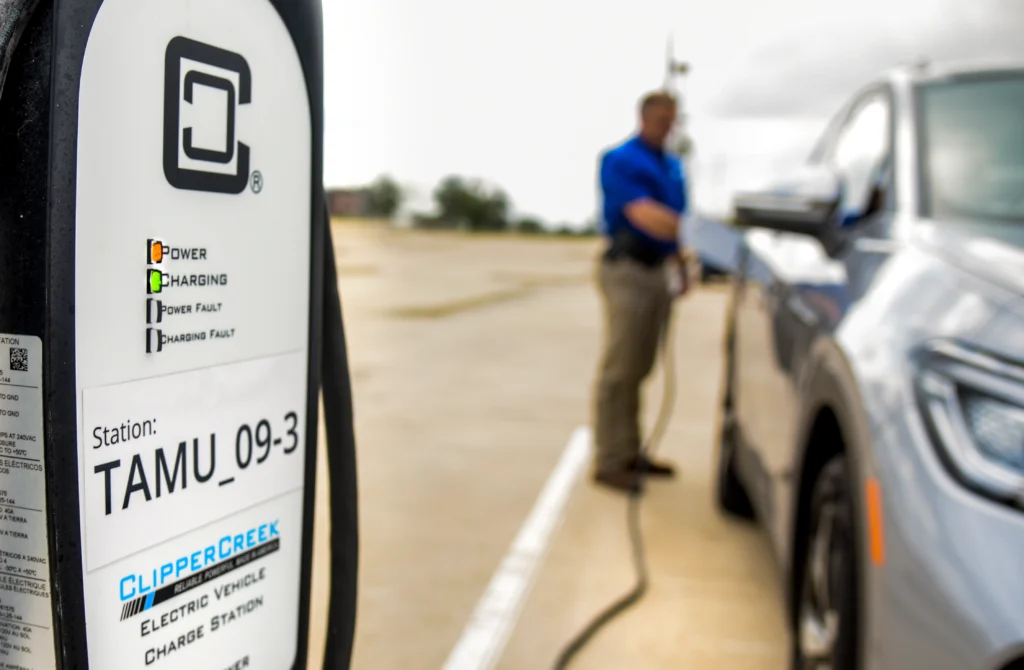 A man in a blue shirt charges an electric vehicle at a ClipperCreek charging station in a parking lot.