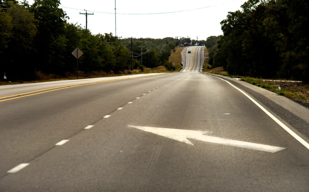 A two-lane road with a white left-turn arrow painted on the surface, lined with trees on both sides, and a vehicle visible in the distance.